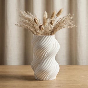 Puede incluir: Un jarr&oacute;n blanco texturizado con un dise&ntilde;o &uacute;nico y retorcido, que contiene hierbas ornamentales secas. El jarr&oacute;n tiene una forma curvil&iacute;nea y est&aacute; sobre una superficie de madera. El fondo es una cortina de tono neutro.