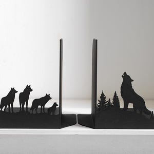 Puede incluir: Un par de sujetalibros de metal negro con siluetas recortadas. Uno muestra una manada de cuatro lobos sobre una cresta. El otro representa a un lobo solitario aullando a la luna en medio de un bosque de pinos con un acabado oscuro.