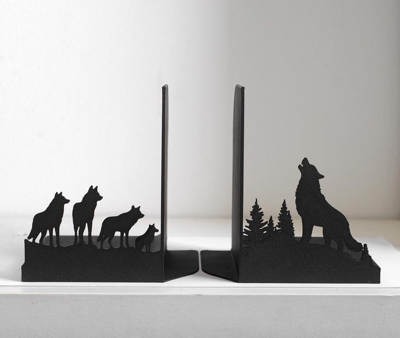 Puede incluir: Un par de sujetalibros de metal negro con siluetas recortadas. Uno muestra una manada de cuatro lobos sobre una cresta. El otro representa a un lobo solitario aullando a la luna en medio de un bosque de pinos con un acabado oscuro.