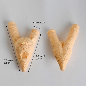 Puede incluir: Dos componentes en forma de V de color beige claro con texturas talladas intrincadas. Una pieza presenta una cabeza de jaguar detallada con la boca abierta. Las dimensiones son 7,3 cm de largo, 6,3 cm de ancho y 1,1 cm de apertura.