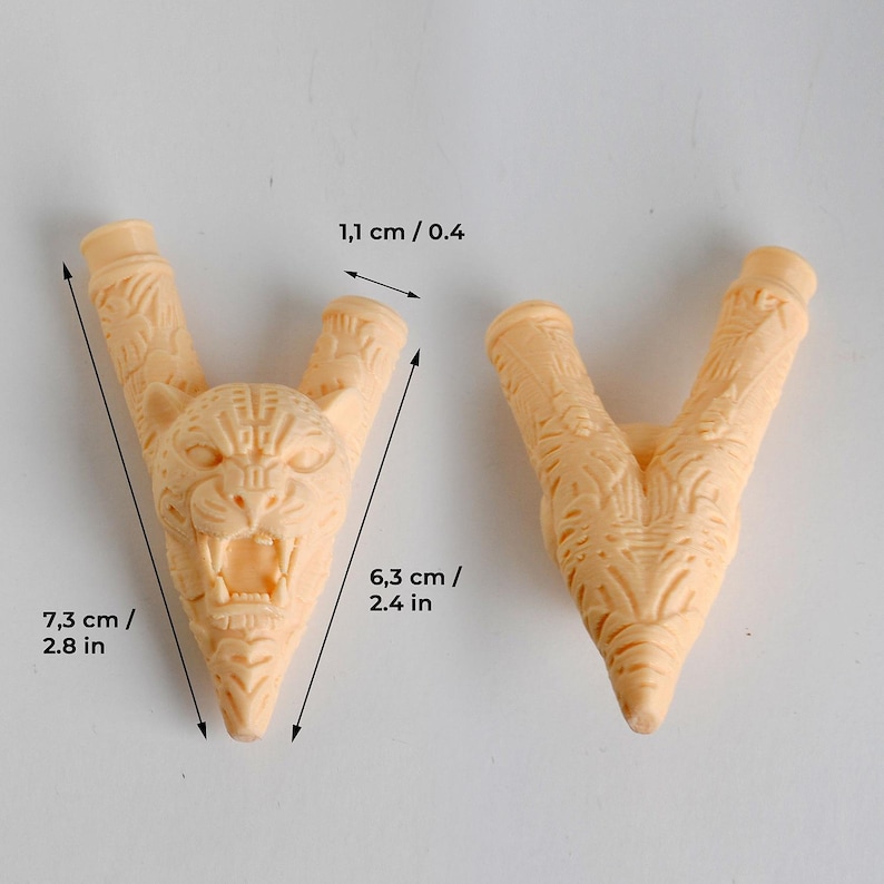 Puede incluir: Dos componentes en forma de V de color beige claro con texturas talladas intrincadas. Una pieza presenta una cabeza de jaguar detallada con la boca abierta. Las dimensiones son 7,3 cm de largo, 6,3 cm de ancho y 1,1 cm de apertura.