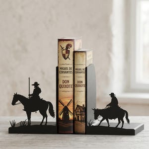 Puede incluir: Un par de sujetalibros de metal negro con siluetas de un jinete a caballo y un jinete en burro, que sostienen dos ejemplares del libro Don Quijote de Miguel de Cervantes, con ilustraciones de un molino de viento y un edificio.