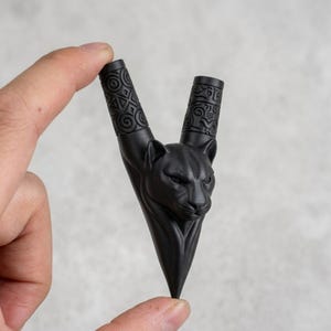 Puede incluir: Una pipa de fumar negra mate en forma de V con una cabeza de pantera esculpida en el centro. Los dos tallos cil&iacute;ndricos superiores est&aacute;n decorados con intrincados patrones geom&eacute;tricos en relieve, mientras la base termina en punta.