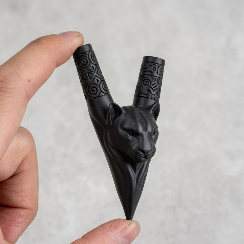 Puede incluir: Una pipa de fumar negra mate en forma de V con una cabeza de pantera esculpida en el centro. Los dos tallos cil&iacute;ndricos superiores est&aacute;n decorados con intrincados patrones geom&eacute;tricos en relieve, mientras la base termina en punta.