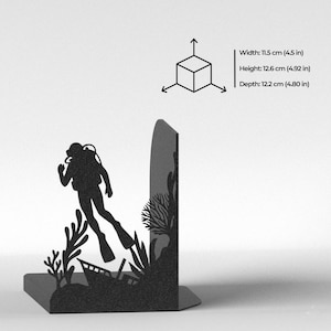 Puede incluir: Un sujetalibros de metal negro con la silueta de un buceador nadando cerca de corales y un naufragio. Las dimensiones son 11,5 cm de ancho, 12,6 cm de alto y 12,2 cm de profundidad, junto a un diagrama de cubo con flechas indicativas.