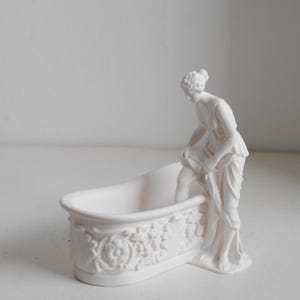 May include: A white matte ceramic figurine featuring a bathtub with ornate floral relief carvings on the exterior. A figure stands beside the tub, lifting the hem of a draped garment while one leg is positioned inside the basin of the bathtub.