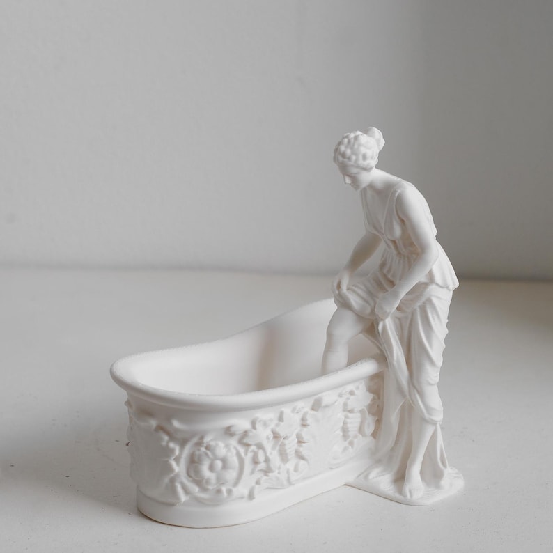 May include: A white matte ceramic figurine featuring a bathtub with ornate floral relief carvings on the exterior. A figure stands beside the tub, lifting the hem of a draped garment while one leg is positioned inside the basin of the bathtub.