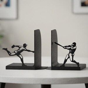 Puede incluir: Un par de sujetalibros de metal negro con siluetas de jugadores de b&eacute;isbol. Un sujetalibros muestra a un lanzador en movimiento de lanzamiento, mientras que el otro muestra a un bateador balanceando un bate. Ambos tienen acabado texturizado.