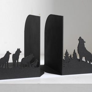 Puede incluir: Un par de sujetalibros de metal negro con siluetas de lobos. Un sujetalibros muestra una manada de cuatro lobos sobre una colina, mientras que el otro muestra a un lobo aullando frente a un bosque de pinos, todo con un acabado texturizado.