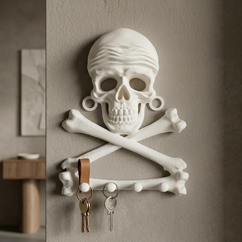 Puede incluir: Un soporte de pared blanco para llaves con forma de calavera y tibias cruzadas. La calavera presenta una textura similar a un pa&ntilde;uelo en la frente y adornos circulares en las orejas. Debajo de los huesos cruzados hay cuatro ganchos.