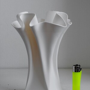 Puede incluir: Un jarr&oacute;n blanco con un acabado mate liso y un borde ondulado y fruncido que se ensancha hacia afuera. El jarr&oacute;n est&aacute; colocado junto a un encendedor desechable de color amarillo ne&oacute;n brillante para escala, resaltando su forma vertical.