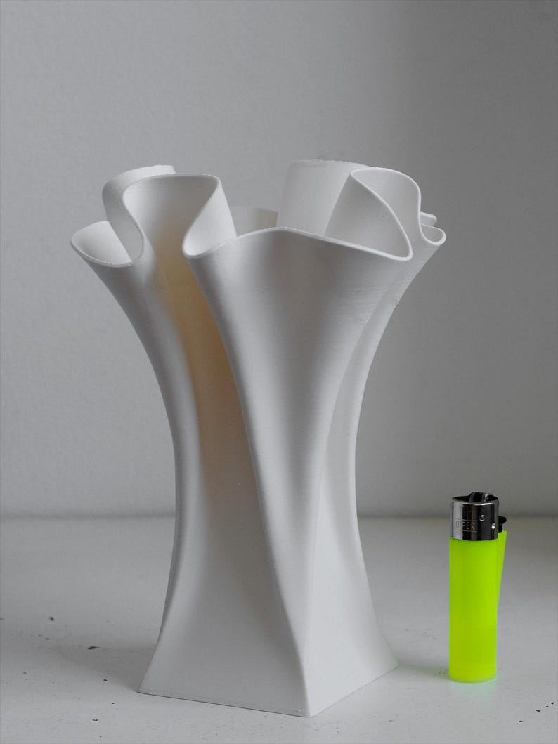 Puede incluir: Un jarr&oacute;n blanco con un acabado mate liso y un borde ondulado y fruncido que se ensancha hacia afuera. El jarr&oacute;n est&aacute; colocado junto a un encendedor desechable de color amarillo ne&oacute;n brillante para escala, resaltando su forma vertical.