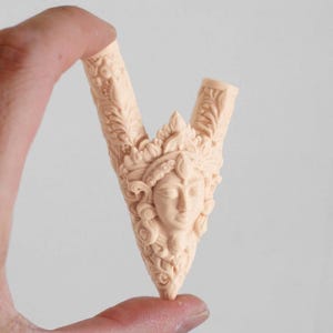 Puede incluir: Un objeto tallado en forma de V de color beige claro, con un relieve central de un rostro humano adornado con un tocado ornamentado. La superficie est&aacute; cubierta de intrincados patrones florales y de hojas que se extienden por ambos brazos de la estructura.