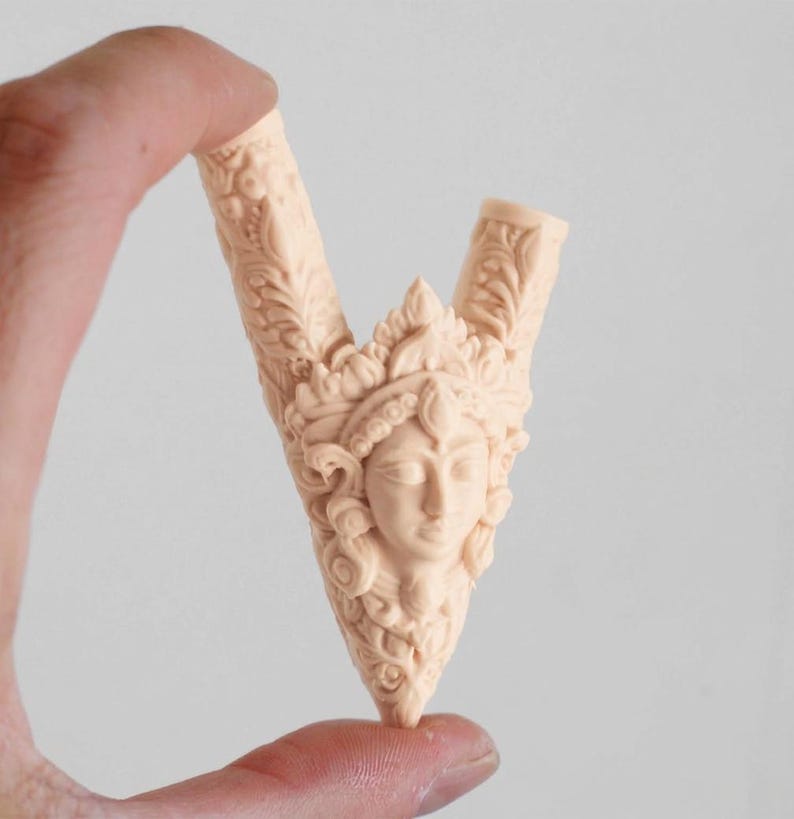 Puede incluir: Un objeto tallado en forma de V de color beige claro, con un relieve central de un rostro humano adornado con un tocado ornamentado. La superficie est&aacute; cubierta de intrincados patrones florales y de hojas que se extienden por ambos brazos de la estructura.
