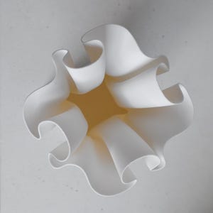 Puede incluir: Un recipiente blanco de acabado mate con un borde ondulado y fruncido que crea una serie de pliegues profundos y curvos. El objeto presenta una base cuadrada que se abre hacia una parte superior amplia con una textura lisa.