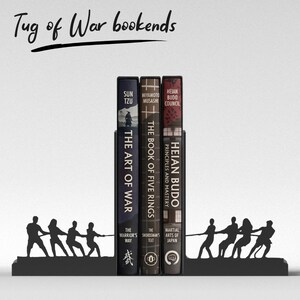 Puede incluir: Soportes para libros de metal negro con un dise&ntilde;o de tira y afloja, sosteniendo tres libros. Los libros se titulan "The Art of War", "The Book of Five Rings" y "Heian Budo". El texto "Tug of War bookends" est&aacute; escrito en la parte superior.