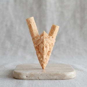 Puede incluir: Una escultura tallada en forma de V de color beige claro que presenta un rostro humano central con los ojos cerrados, rodeado de intrincados patrones en relieve de flores y hojas. El objeto tiene dos aberturas cil&iacute;ndricas huecas arriba.