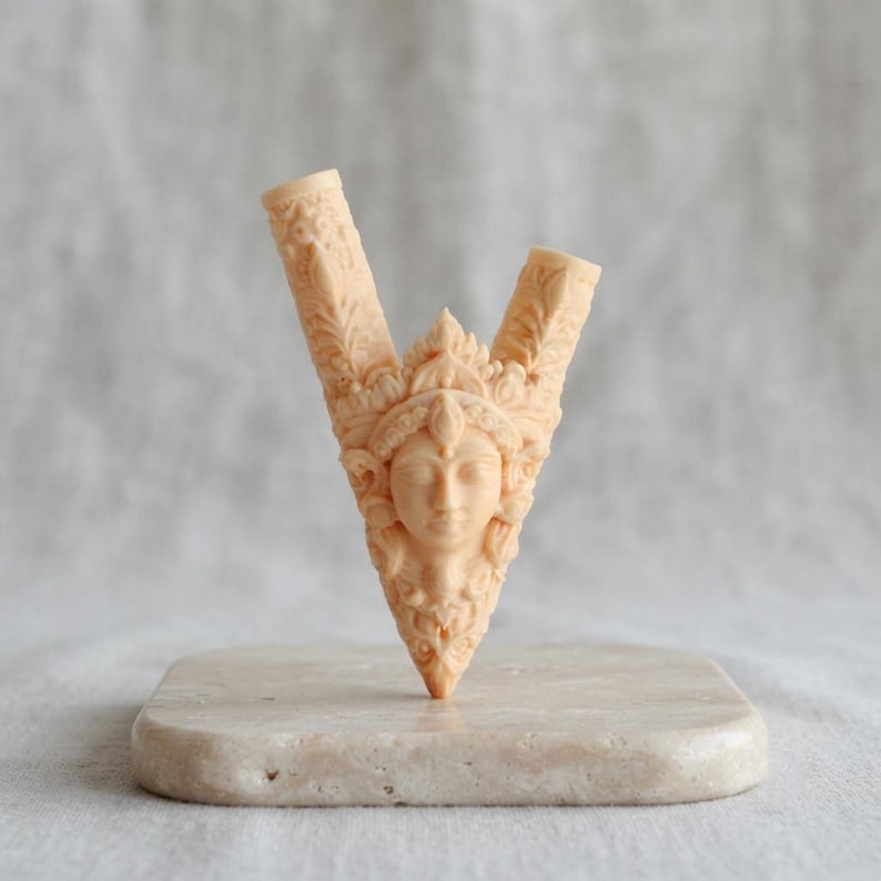 Puede incluir: Una escultura tallada en forma de V de color beige claro que presenta un rostro humano central con los ojos cerrados, rodeado de intrincados patrones en relieve de flores y hojas. El objeto tiene dos aberturas cil&iacute;ndricas huecas arriba.
