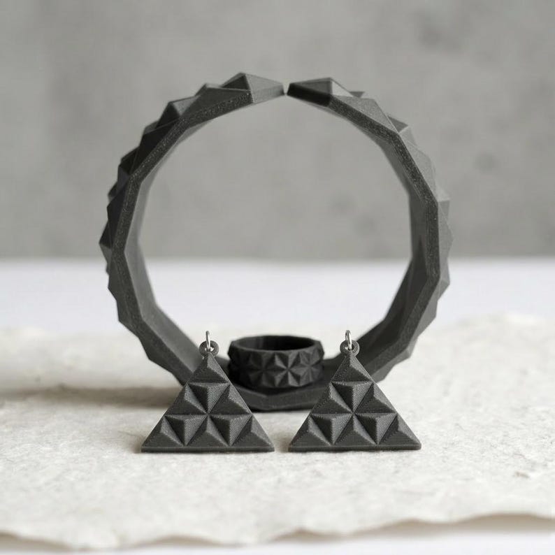 May include: A set of geometric jewelry in a dark gray color. The set includes a ring, a pair of triangular earrings, and a large, open circular pendant. The earrings and ring have a repeating triangular pattern. The jewelry is displayed on a textured, off-white surface.