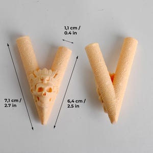Op de afbeelding: Twee licht perzikkleurige V-vormige objecten met ingewikkelde reli&euml;fpatronen en een centrale schedelfiguur met een hoofdtooi. De aangegeven afmetingen zijn 1,1 cm breed, met hoogtes van respectievelijk 7,1 cm en 6,4 cm.
