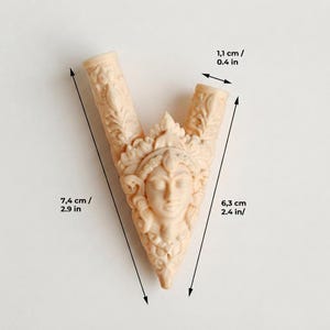 Puede incluir: Un objeto tallado en forma de V de color beige claro que presenta un rostro central con tocado ornamentado y detalles florales. El objeto mide 7,4 cm de largo total, 6,3 cm en el lado derecho y tiene una abertura superior de 1,1 cm.