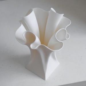 Puede incluir: Un florero blanco mate con base cuadrada que se transforma en un borde ancho, ondulado y con volantes. La estructura presenta curvas profundas y fluidas que crean una apariencia din&aacute;mica y plisada, extendi&eacute;ndose desde el cuello hasta la boca.