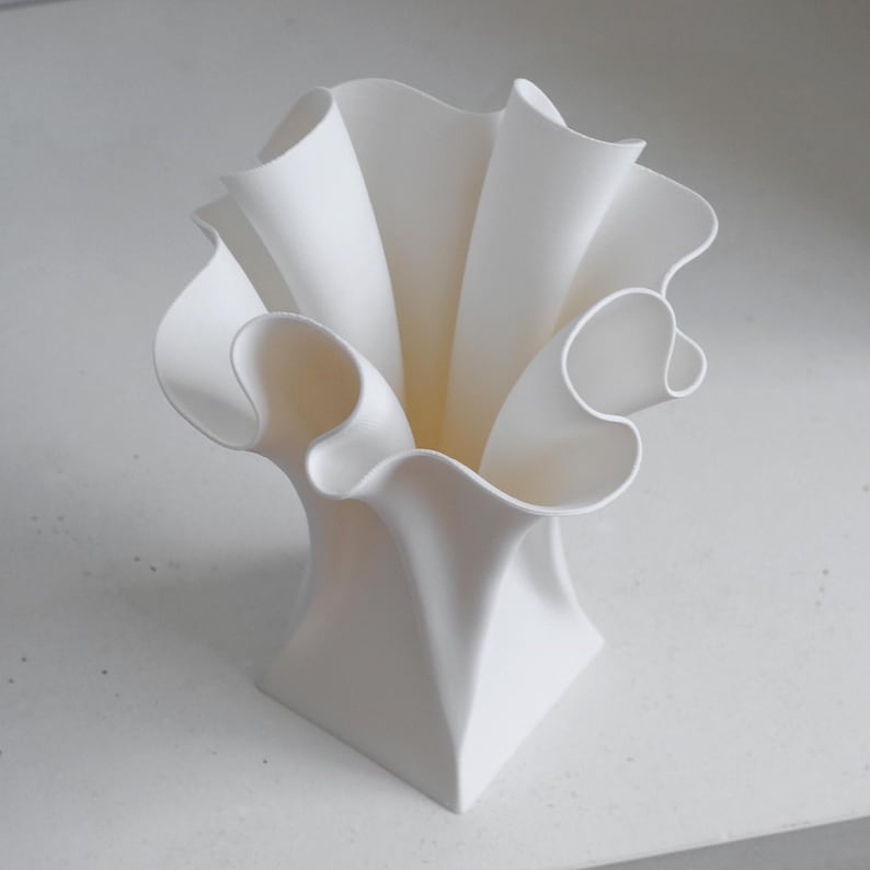 Puede incluir: Un florero blanco mate con base cuadrada que se transforma en un borde ancho, ondulado y con volantes. La estructura presenta curvas profundas y fluidas que crean una apariencia din&aacute;mica y plisada, extendi&eacute;ndose desde el cuello hasta la boca.