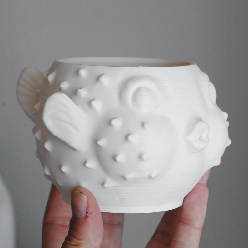 Puede incluir: Una taza de cer&aacute;mica blanca mate con forma de pez globo. El exterior presenta una superficie texturizada con espinas en relieve, aletas moldeadas en los laterales, un ojo circular y un detalle de boca fruncida. El recipiente es redondeado.