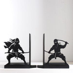 Puede incluir: Un par de sujetalibros de metal negro con siluetas de ninjas en posturas de combate din&aacute;micas. Cada sujetalibros muestra una figura ninja sosteniendo una espada, con un ninja en cuclillas y el otro en una postura defensiva, hechos de metal negro.