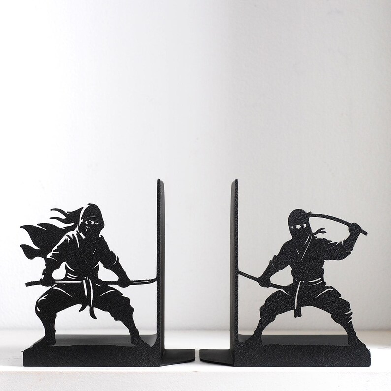Puede incluir: Un par de sujetalibros de metal negro con siluetas de ninjas en posturas de combate din&aacute;micas. Cada sujetalibros muestra una figura ninja sosteniendo una espada, con un ninja en cuclillas y el otro en una postura defensiva, hechos de metal negro.