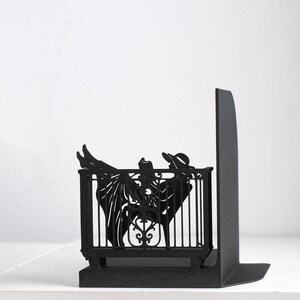 Puede incluir: Un sujetalibros de metal negro mate con la silueta de una figura con sombrero de ala ancha, reclinada en una silla detr&aacute;s de una barandilla decorativa. La figura sostiene un libro abierto, con las piernas extendidas hacia arriba.