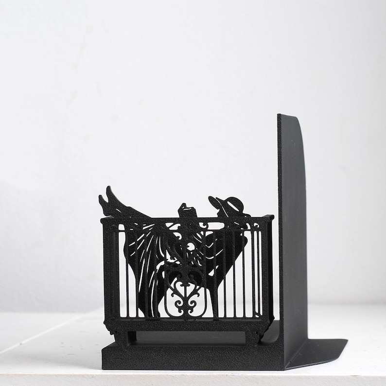 Puede incluir: Un sujetalibros de metal negro mate con la silueta de una figura con sombrero de ala ancha, reclinada en una silla detr&aacute;s de una barandilla decorativa. La figura sostiene un libro abierto, con las piernas extendidas hacia arriba.