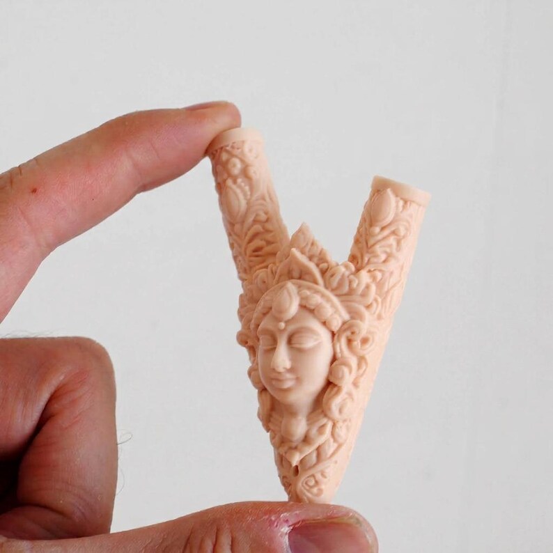 Puede incluir: Una escultura en forma de V de color melocot&oacute;n claro que presenta un relieve central de un rostro humano rodeado de intrincadas tallas florales y volutas. El objeto tiene dos puntas que se extienden hacia arriba y un acabado mate liso.