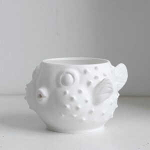 Puede incluir: Una taza de cer&aacute;mica blanca con forma de pez globo, que presenta un cuerpo redondeado con espinas texturizadas en relieve en la superficie, una peque&ntilde;a boca protuberante y aletas esculpidas en los lados, con una parte superior abierta.