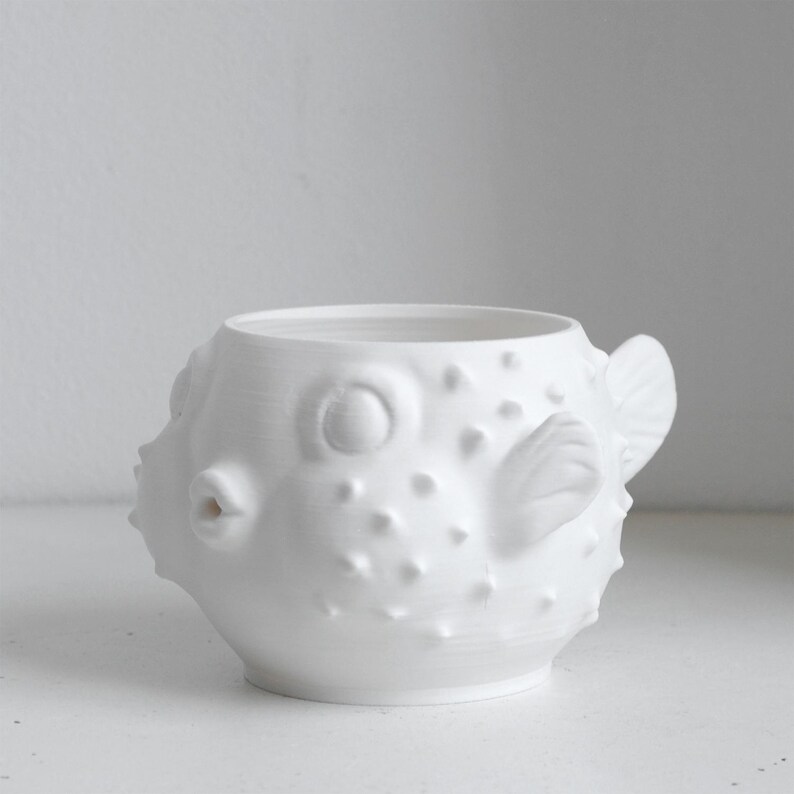 Puede incluir: Una taza de cer&aacute;mica blanca con forma de pez globo, que presenta un cuerpo redondeado con espinas texturizadas en relieve en la superficie, una peque&ntilde;a boca protuberante y aletas esculpidas en los lados, con una parte superior abierta.