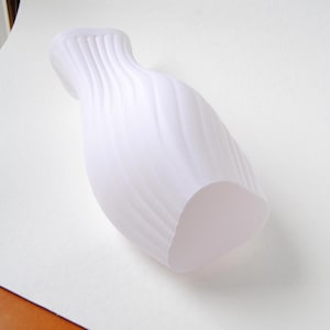Puede incluir: Un florero blanco mate que presenta una textura acanalada vertical distintiva que se espiraliza ligeramente a lo largo de su cuerpo. El recipiente tiene un cuello estrecho que se ensancha hacia la abertura superior y una base redondeada.