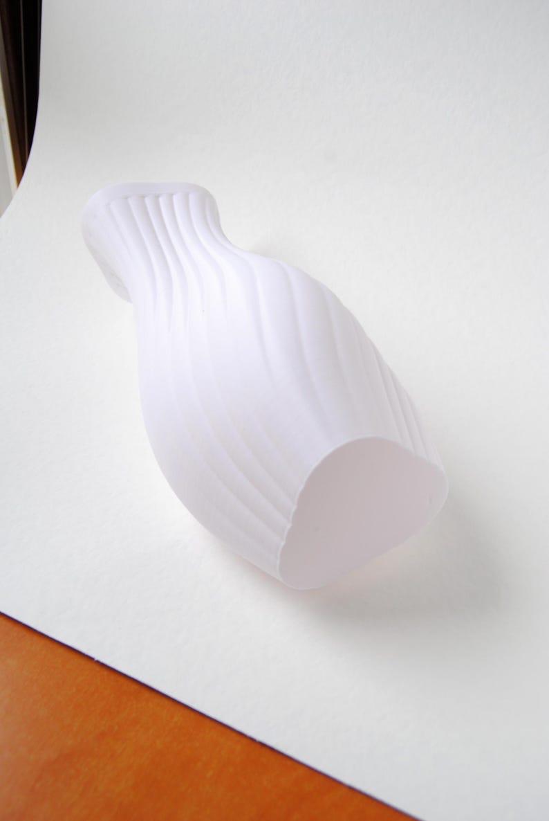 Puede incluir: Un florero blanco mate que presenta una textura acanalada vertical distintiva que se espiraliza ligeramente a lo largo de su cuerpo. El recipiente tiene un cuello estrecho que se ensancha hacia la abertura superior y una base redondeada.