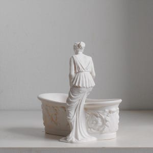May include: A matte white ceramic planter shaped like a bathtub with a sculpted figure standing at the edge. The exterior of the tub features detailed relief carvings of floral patterns and leaves, while the figure wears a draped garment.