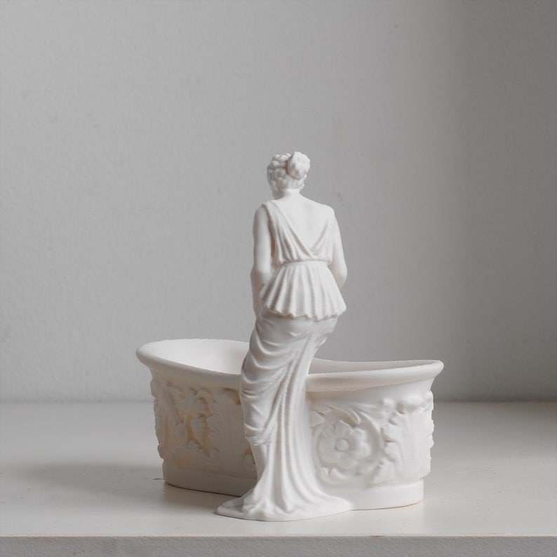 May include: A matte white ceramic planter shaped like a bathtub with a sculpted figure standing at the edge. The exterior of the tub features detailed relief carvings of floral patterns and leaves, while the figure wears a draped garment.