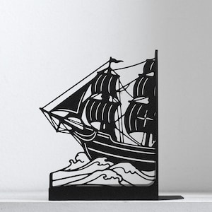 Puede incluir: Un sujetalibros de metal negro con la silueta de un velero con varios m&aacute;stiles y velas navegando a trav&eacute;s de olas estilizadas. El dise&ntilde;o est&aacute; cortado de una sola pieza de metal plano, creando un perfil n&iacute;tido y de alto contraste.