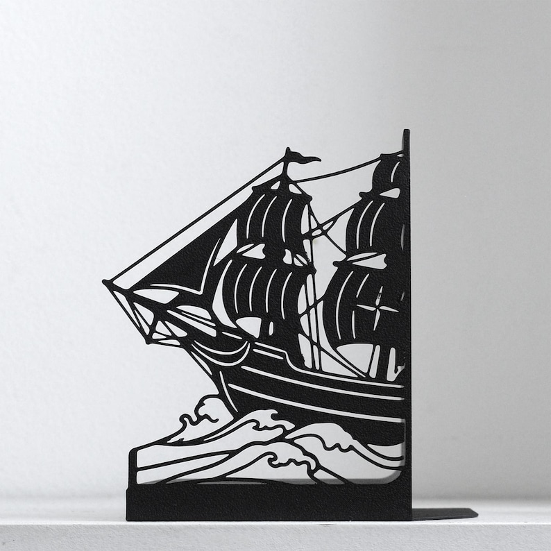 Puede incluir: Un sujetalibros de metal negro con la silueta de un velero con varios m&aacute;stiles y velas navegando a trav&eacute;s de olas estilizadas. El dise&ntilde;o est&aacute; cortado de una sola pieza de metal plano, creando un perfil n&iacute;tido y de alto contraste.