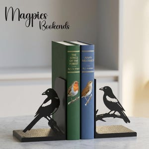 Puede incluir: Un par de sujetalibros de metal negro con siluetas de urracas posadas en ramas. Los sujetalibros sostienen dos libros de tapa dura titulados The Songs of the Forest y Avian Melodies, que presentan ilustraciones de aves en sus lomos.
