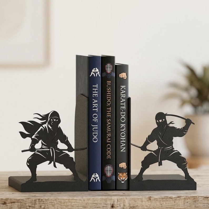 Puede incluir: Un par de sujetalibros de metal negro con siluetas de ninjas en posturas de combate din&aacute;micas, cada uno sosteniendo una espada. Los sujetalibros sostienen tres libros titulados The Art of Judo, Bushido: The Samurai Code y Karate-Do Kyohan.