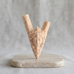 Puede incluir: Un objeto tallado en forma de V de color beige claro, con un rostro humano central rodeado de intrincados relieves de flores y hojas. El objeto tiene dos aberturas cil&iacute;ndricas huecas en la parte superior y se asienta sobre una base de piedra.