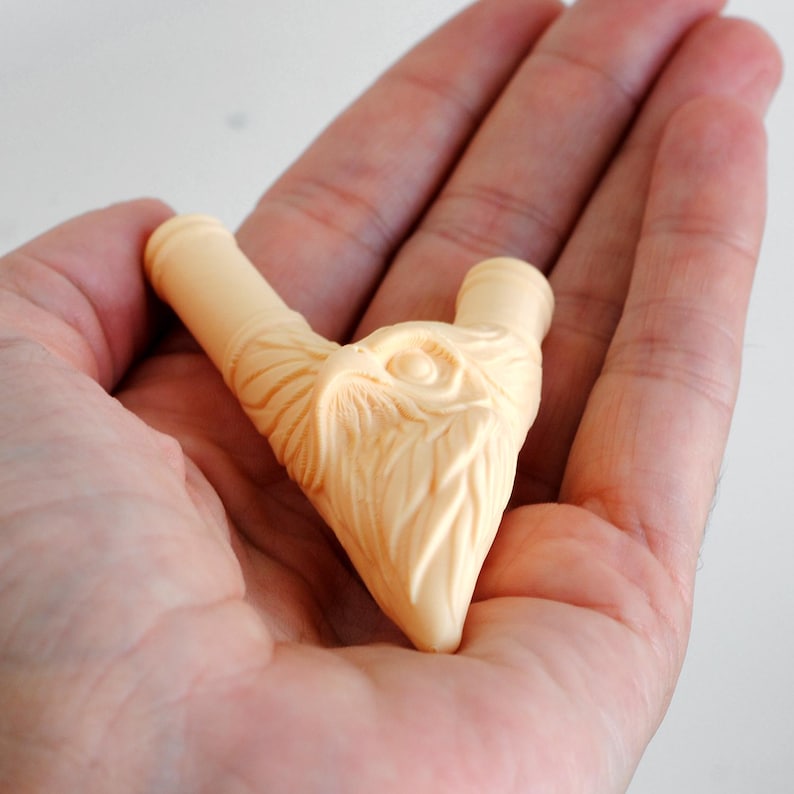 Puede incluir: Un objeto tallado de color beige claro que presenta la cabeza detallada de un b&uacute;ho con plumas texturizadas y dos aberturas cil&iacute;ndricas en la parte superior. La pieza tiene un acabado liso y muestra rasgos faciales intrincados.