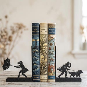 Puede incluir: Tres libros de tapa dura titulados The Odyssey, Plant Myths y Mapping the Unknown se sostienen entre dos sujetalibros de metal negro. El sujetalibros izquierdo muestra la silueta de una persona con paraguas y el derecho a una persona paseando un perro.