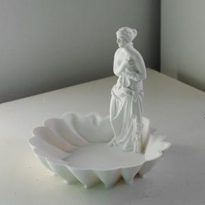 Puede incluir: Un cuenco decorativo de color blanco mate con un borde festoneado similar a una concha y una figura central erguida de una silueta cl&aacute;sica drapeada en tela. La escultura est&aacute; integrada en el cuenco, creando un objeto blanco unificado.