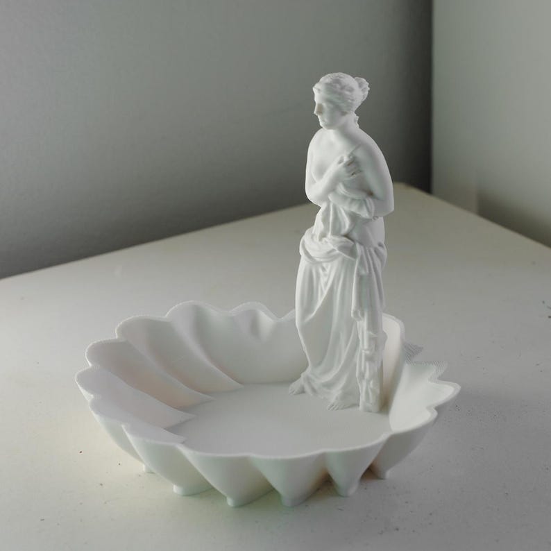 Puede incluir: Un cuenco decorativo de color blanco mate con un borde festoneado similar a una concha y una figura central erguida de una silueta cl&aacute;sica drapeada en tela. La escultura est&aacute; integrada en el cuenco, creando un objeto blanco unificado.