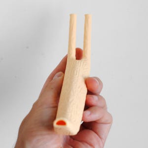 Puede incluir: Un silbato de pl&aacute;stico de color melocot&oacute;n claro con un patr&oacute;n geom&eacute;trico texturizado en el cuerpo principal y dos tubos verticales largos y delgados que se extienden desde la parte superior. La boquilla tiene una abertura naranja.
