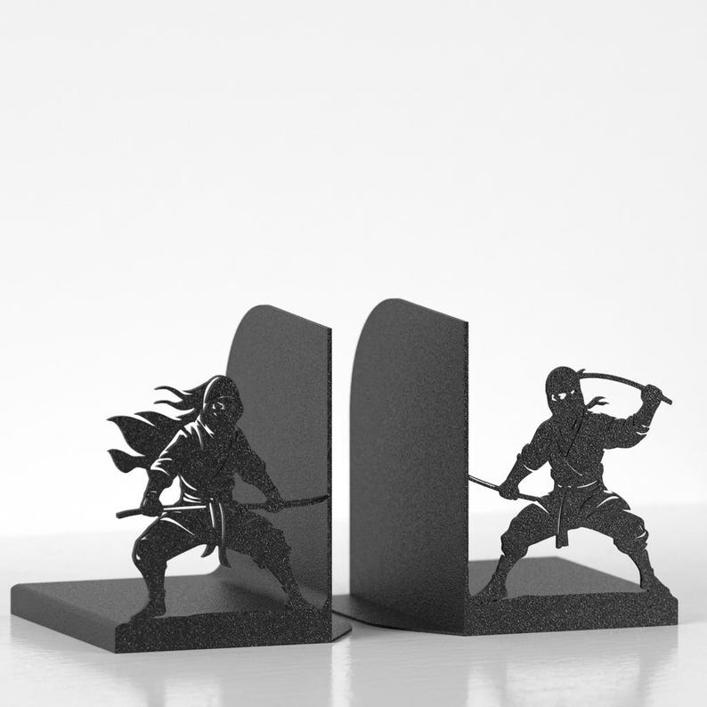 Puede incluir: Un par de sujetalibros de metal negro con siluetas cortadas con l&aacute;ser de ninjas en posturas de combate din&aacute;micas. Cada sujetalibros muestra una figura ninja sosteniendo una espada, con una figura avanzando y la otra en una postura defensiva.